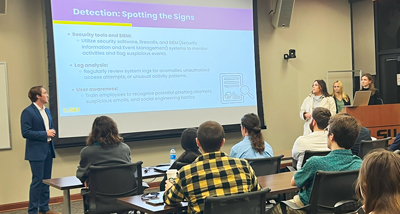 Students present about cybersecurity, they refer to a slide and gesture as the crowd listens.