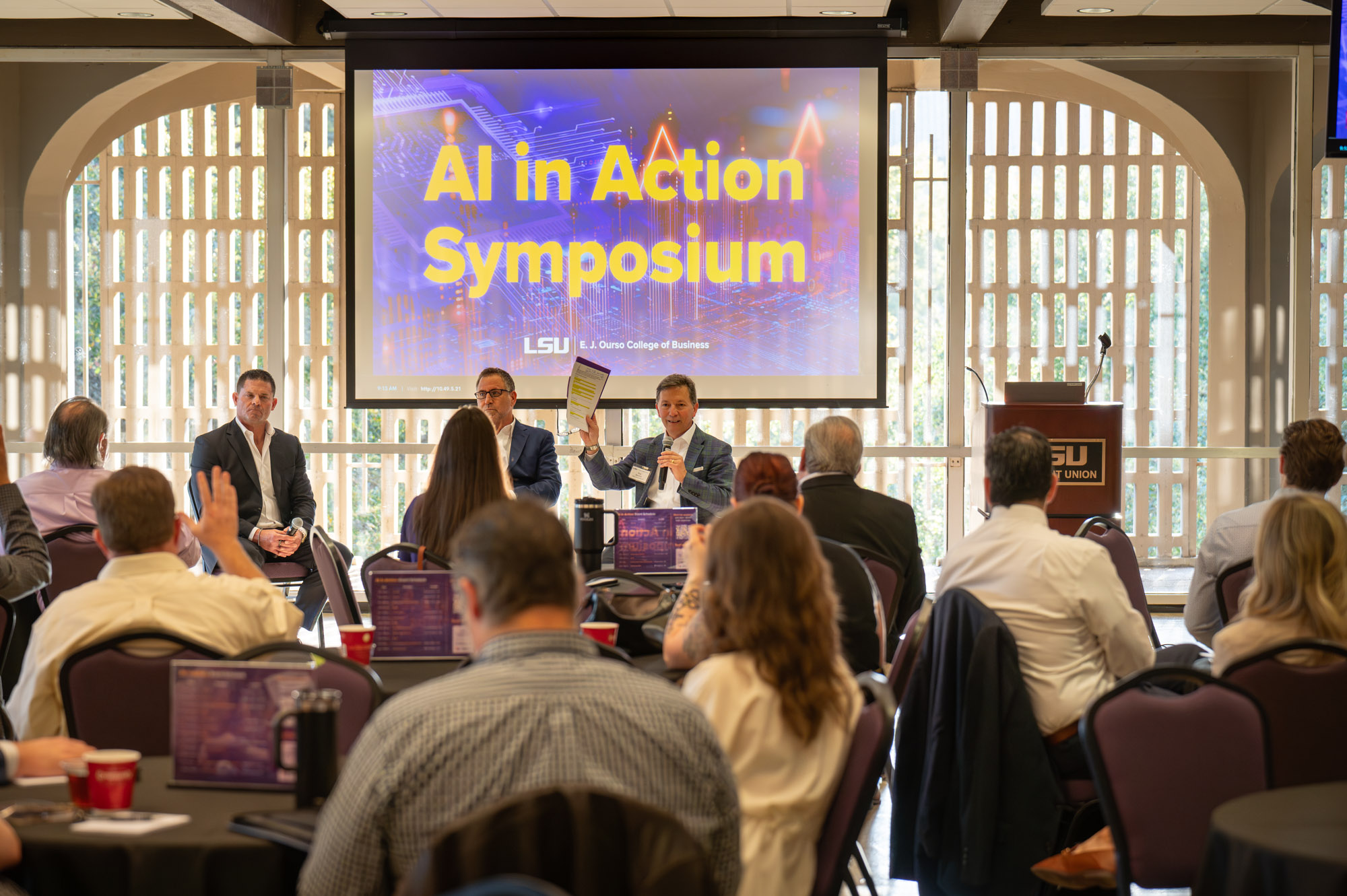 A group of men address the packed room in the LSU Student Union. The moderator holds up a pad of paper and there is a raised hand in the audience.